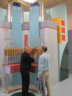 In this wooden engineering mock-up, module 3L is seen in crosssection, the upper part of the toroidal field coils is represented at the back with the precompression ring fixture in orange, and the vertical tie-plates (part of the central solenoid precompression structure) are represented in light blue. Charles Lyraud and Neil Mitchell from the ITER Organization talk over insulating breaks. (Click to view larger version...)
