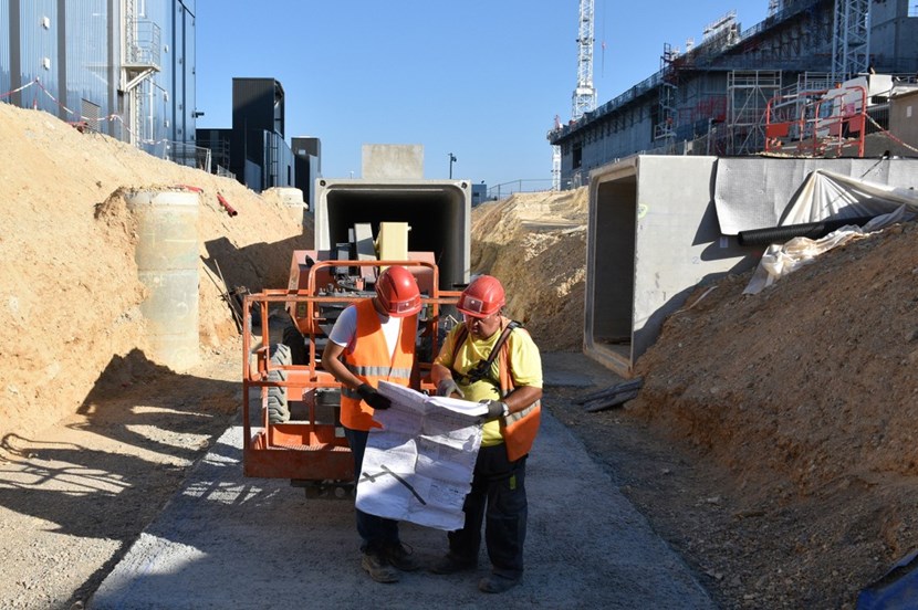A service tunnel opens up at a junction where workers install pre-fabricated elements to connect the galleries coming from three directions. The opening offers a view of the Tokamak Complex on the right and the cryoplant on the left. (Click to view larger version...)